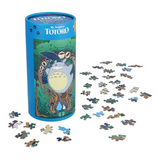 A cylindrical blue puzzle box featuring Totoro from My Neighbor Totoro, with many small  puzzle pieces scattered around it on a white background.