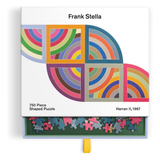 A 750-piece shaped puzzle box by Frank Stella, shown open with the puzzle pieces sliding out. The box features a colorful geometric design inspired by Harran II, 1967, and the puzzle pieces below are multicolored.
