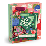 A boxed jigsaw puzzle package titled "Mahjong Social" shown at a slight angle on a white background. The box features colorful illustrated artwork of a mahjong table scene with tiles, flowers, teapot, cups, and furniture.