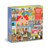 A 300-piece jigsaw puzzle box titled 'Main Street Morning' by The Puzzle Factory. The box features a colorful, highly detailed illustration of a quaint village street with brick bakeries, colorful houses, and people with bicycles.