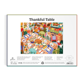 Front cover of a jigsaw puzzle package titled "Thankful Table," showing a colorful illustrated scene of a table laden with food and holiday decorations. The package includes product text, a barcode, and puzzle piece graphics on a white background.