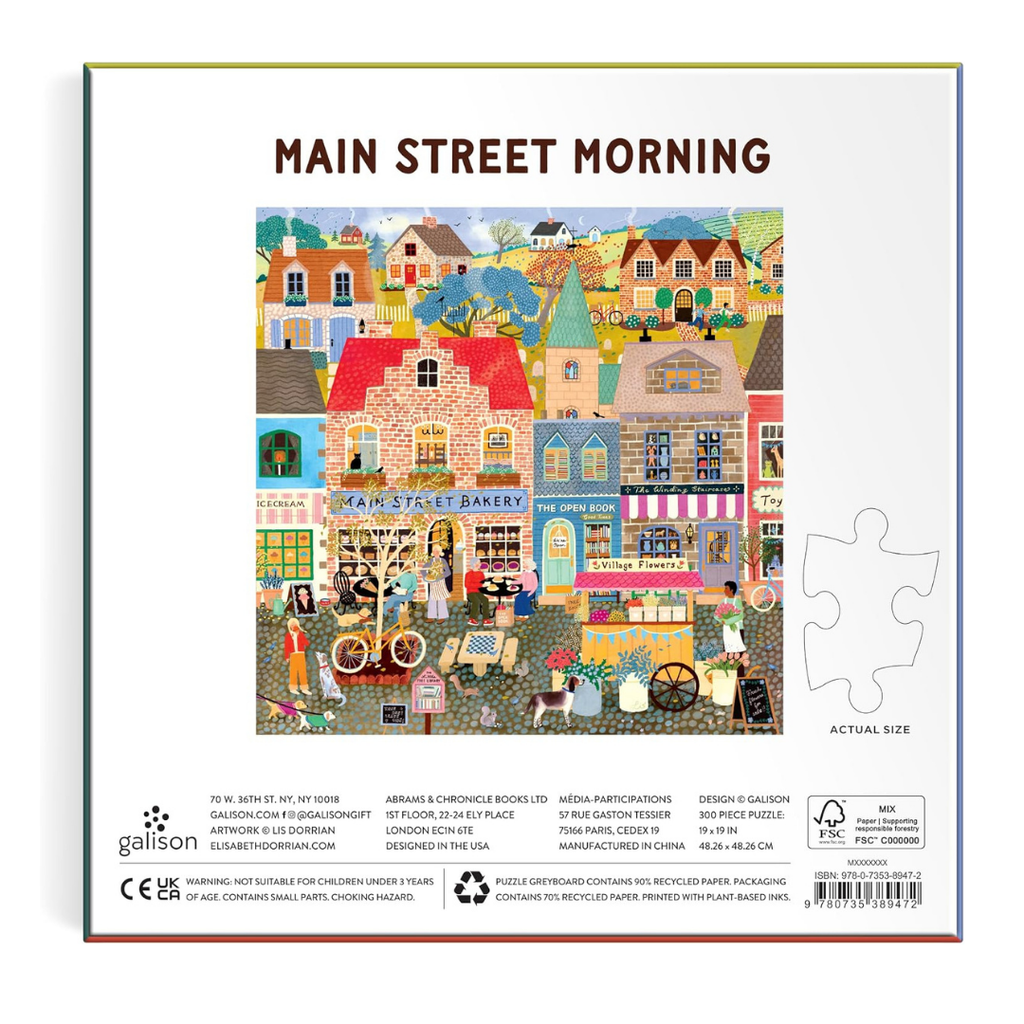 Back of a Galison 'Main Street Morning' jigsaw puzzle box, showing a colorful illustrated town scene with houses, bakery, people, and street details, plus a white puzzle-piece outline icon and product information at the bottom.