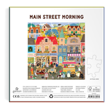 Back of a Galison 'Main Street Morning' jigsaw puzzle box, showing a colorful illustrated town scene with houses, bakery, people, and street details, plus a white puzzle-piece outline icon and product information at the bottom.
