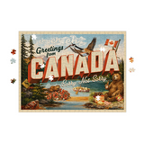 A completed jigsaw puzzle featuring a vintage-style Canadian travel poster with large red 'CANADA' lettering, a bird, a bear, trees, and the text 'Greetings from Canada' and 'Sorry Not Sorry', shown on a white background with a few loose puzzle pieces around it.