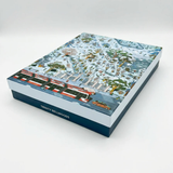 A rectangular puzzle box with a detailed winter landscape illustration on the lid, featuring snow-covered trees, buildings, and a red-and-white train. The side of the box is dark blue with the text 'TRINITY BELLWOODS' in white.