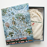 A winter-themed illustrated jigsaw puzzle box shown open, with a cream drawstring pouch inside and a printed cover featuring a snowy city scene and transit vehicles. The pouch has green text reading 'pieces over pixels.