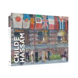 Ajigsaw puzzle box shown at a slight angle on a white background. The front features an impressionistic painting of a street scene with colorful flags and pedestrians, and the spine/cover text reads CHILDE HASSAM.