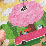 A colorful children's jigsaw puzzle showing a pink car with cartoon characters, set against green hills and a large pink heart-shaped tree, photographed on a wooden surface with flowers in the foreground.