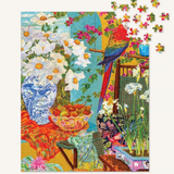 A colorful completed jigsaw puzzle showing a vibrant still life scene with white daisies in a blue-and-white vase, a red-and-blue parrot, flowers, fruit, and furniture, with several loose puzzle pieces floating at the top right on a light neutral background.