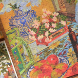 A colorful jigsaw puzzle featuring a still life painting with a blue patterned vase, pink flowers, green leaves, and red apples, photographed close up on a wooden surface.