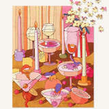 A colorful illustrated jigsaw puzzle showing whimsical cartoon characters in cocktail glasses, candles, flowers, and fruit on a warm-toned background. The puzzle pieces are scattered at the top right, and the puzzle itself is photographed against a plain light background.