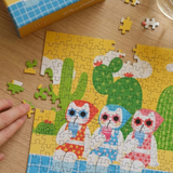 A colorful jigsaw puzzle showing three cartoon cats in swimwear among cacti, partially assembled on a wooden table with a hand visible at the left edge and a few loose puzzle pieces scattered around. The box is partly visible in the top left.