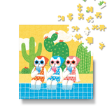 A square jigsaw puzzle featuring three cartoon cats sitting by a blue pool with cacti in the background, shown on a white surface with several loose puzzle pieces floating above.