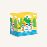 A cube-shaped jigsaw puzzle box featuring a colorful desert illustration with cacti, clouds, and three stylized characters sitting by a blue tiled pool. The box is photographed on a clean, light off-white background.
