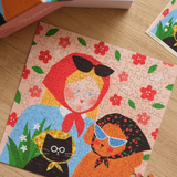 A colorful children's jigsaw puzzle featuring an illustration of a girl in a red bonnet, a black cat in a yellow bonnet, and a dog wearing sunglasses, set on a light wood tabletop.
