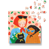 A completed jigsaw puzzle showing a whimsical illustration of a woman in a red headscarf with sunglasses, a woman in a black floral headscarf with blue sunglasses, a black cat in a yellow bonnet, and flowers on a peach background. Several loose puzzle pieces are scattered above on a white background.