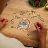 A person's hands arranging puzzle pieces on a light wood table, completing a colorful stained-glass style jigsaw puzzle. A green holiday wreath and a lit candle are visible in the background.