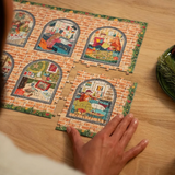 A person's hand is placing a puzzle piece onto a set of four jigsaw puzzles on a light wood table. The puzzles feature colorful, whimsical illustrations of cozy indoor scenes framed by brick arches with green foliage borders.