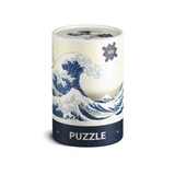 A cylindrical puzzle tin featuring a print of Katsushika Hokusai's Great Wave off Kanagawa. The design shows a large blue and white wave with Mount Fuji in the background, a cream-colored sky, and a dark navy blue band at the bottom with the word 'PUZZLE' in white.