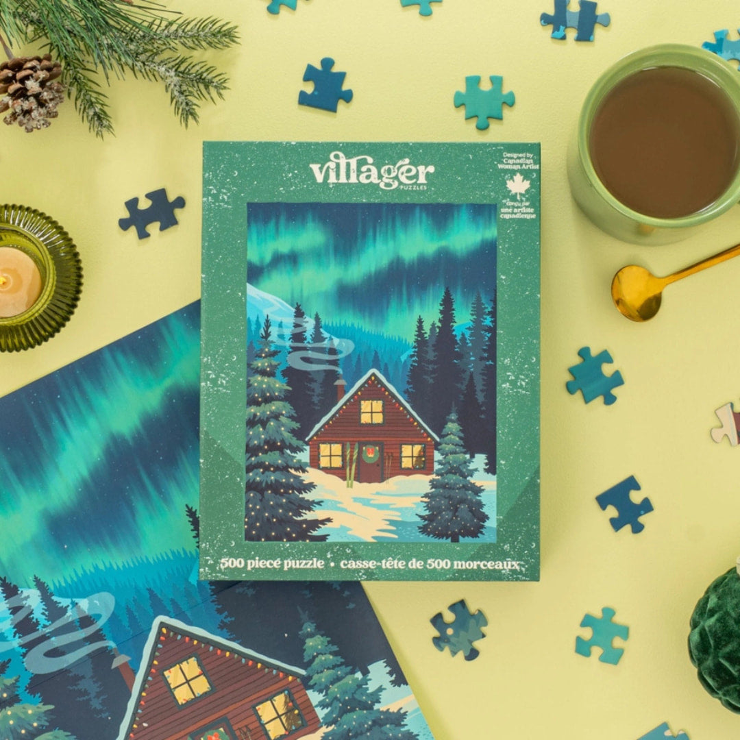 Villager puzzle box with Northern Lights design surrounded by puzzle pieces, a mug of coffee, and decorative items on a yellow background.