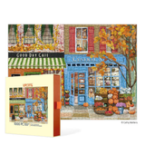 A 1000-piece jigsaw puzzle box shown in front of the completed puzzle, both featuring a colorful autumn street scene with storefronts, pumpkins, and fall foliage. The box reads 'Werkshoppe 1000 PC PUZZLE' and the completed puzzle has 'Good Day Cafe' and 'General Store' visible.
