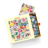 A 1000-piece jigsaw puzzle box by Werkshoppe, featuring a colorful beach umbrella and people pattern on the cover, shown open with loose puzzle pieces visible inside a gold tray on a white background.