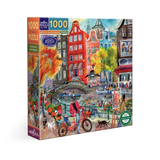 A 1000-piece jigsaw puzzle box by eeBoo Puzzles, featuring a colorful illustration of a canal-side town with red and yellow buildings, a bridge, boats, and a person riding a red bicycle in the foreground.