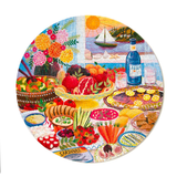 A round jigsaw puzzle featuring a vibrant Mediterranean table setting with an assortment of fresh fruits, cheeses, cured meats, and a bottle of Agua de Marina, with a scenic coastal sunset in the background.