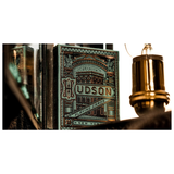A vintage-style playing card deck box labeled HUDSON Premium Playing Cards, shown in a moody close-up with warm lighting and a brass lamp visible on the right.