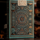 An ornate playing card deck box with intricate geometric and celestial artwork in teal, gold, and black, standing upright against a dark blurred background.