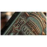 Close-up of an embossed deck of cards cover with ornate architectural detailing in metallic copper over a muted teal-green background.
