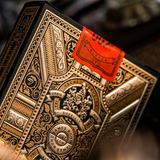 An ornate black and gold playing card deck box with intricate embossed decorative patterns and a bright orange seal attached near the top, photographed in a close-up shot against a dark blurred background.