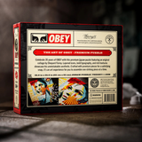 The back of a boxed jigsaw puzzle, branded OBEY and theory 11, shown standing upright on a dark textured surface. The package is mostly black with a cream back panel, red accents, and puzzle artwork panels along the bottom.