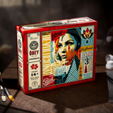 A red Obey brand 'Revolution' 3-piece jigsaw puzzle box displayed on a dark wooden surface with paintbrushes and a paint bottle nearby, indicating a creative or artistic theme.