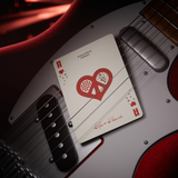 A close-up of a playing card with a red heart design and the text 'England's Finest' and 'Ace of Hearts' resting on the body of a red and silver electric guitar.