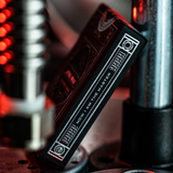 A close-up of a black deck of playing cards with silver engraved text reading 'NOW I AM THE MASTER' and circular emblem details, set against a dark, blurred industrial background with red light reflections.