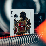 A close-up of a playing card showing the King of Spades with a stylized Darth Vader-like figure in black, red, and gold, set against a dark blurred background with bright orange light.