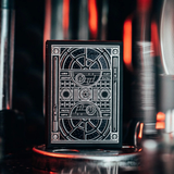 A black playing card deck with intricate white sci-fi themed line art and text, standing on a reflective surface with a dark, blurred background featuring warm red-orange light streaks.