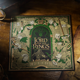A square jigsaw puzzle featuring The Lord of the Rings artwork, photographed on a rustic wooden surface. The front shows illustrated characters and the title 'The Lord of the Rings', in green, gold, and dark tones.