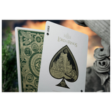 A close-up of playing cards featuring Lord of the Rings artwork, with the Ace of Spades front card in front and the back of another card visible behind it. The cards show green and gold fantasy-themed designs on a blurred stone-and-nature background.
