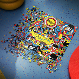 A colorful jigsaw puzzle featuring The Beatles and the Yellow Submarine, shown partially assembled on a blue speckled surface with loose puzzle pieces scattered around it.