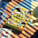 A boxed card game titled "Yellow Submarine" by Beatles lies on a wooden tabletop with sunlight and shadows. The box is brightly illustrated in yellow, blue, black, and white, and cards are partially visible around it.