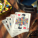 A fan of Beatles-themed playing cards on a wooden tabletop, with a Beatles-themed box partially visible at left, a black leather belt at top right, and audio equipment props around the scene.