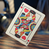 A standard playing card, specifically the Queen of Hearts, shown floating at an angle against a blurred background of a wooden table and dark objects.