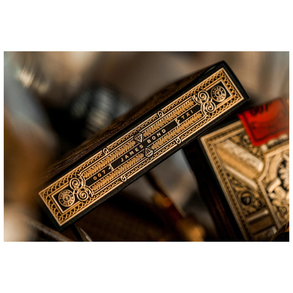 A close-up of a deck of playing cards showing the spine with ornate gold and black detailing, alongside part of the back of another card deck in a warm, blurred setting.