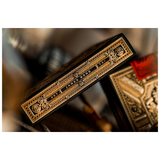 A close-up of a deck of playing cards showing the spine with ornate gold and black detailing, alongside part of the back of another card deck in a warm, blurred setting.