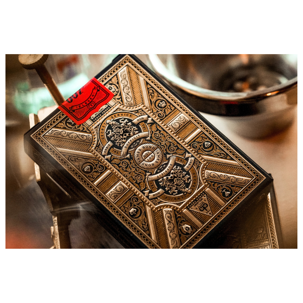 An ornate decorative card deck box with embossed metallic gold and silver patterns, featuring a red and black warning label on the top left, photographed on a reflective surface with a blurred metal bowl in the background.