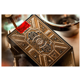 An ornate decorative card deck box with embossed metallic gold and silver patterns, featuring a red and black warning label on the top left, photographed on a reflective surface with a blurred metal bowl in the background.